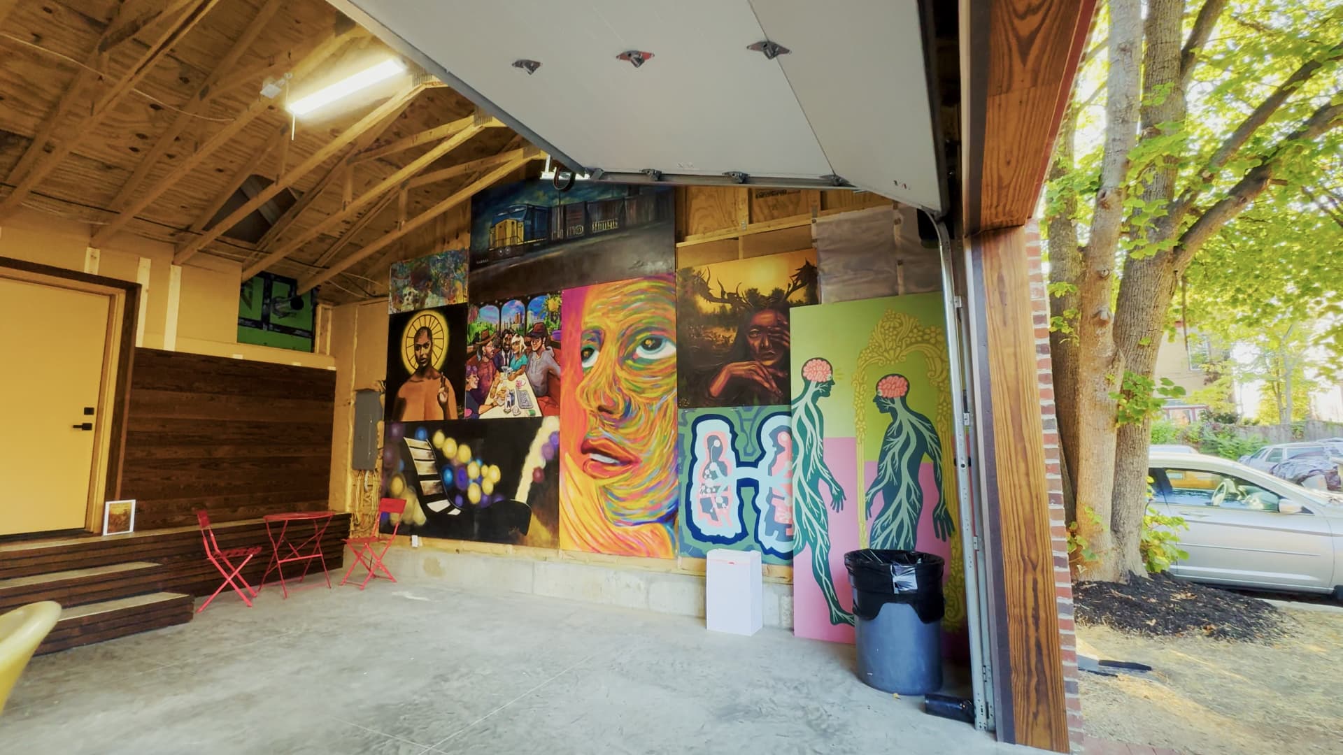 Open garage bay at 990 Oak with the overhead door raised, exposed timber roof framing, a polished concrete floor, and a wall of large painted canvases used as a gallery and flex space